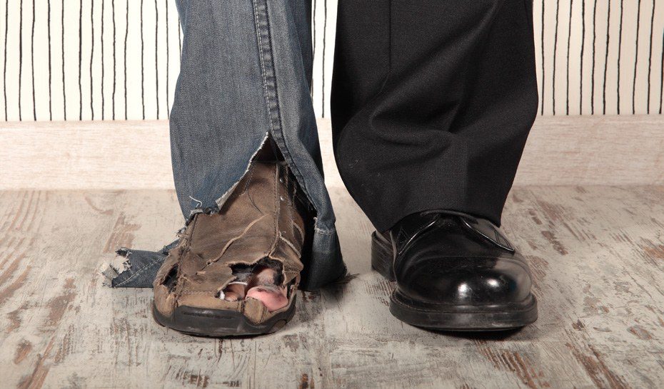 Zwei Beine nebeneinander: links eine abgewetzte Jeans und ein zerrissener, löchriger Lederschuh, aus dem die Zehen schauen, rechts eine schwarze Anzughose und ein glänzender schwarzer Schuh.